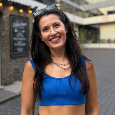 Tuğba, a woman with dark hair wearing a blue top and layered necklaces, smiling on a city street with buildings behind her.